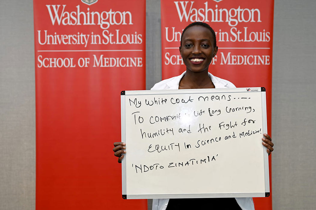 A young Black woman wearing a white physicians coat holds a handwritten sign that says: "My white coat means ... to commit to lifelong learning, humility and the fight for equity in science and medicine! 'Ndoto Zinatimia'"